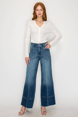 A woman modeling a white long-sleeve button-up top and high-waisted, wide-leg blue denim jeans with contrast stitching and cuffed hems, standing against a plain white studio background.