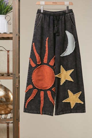 Black pants with sun, moon, and star designs hanging on a wooden hanger against a neutral wall.