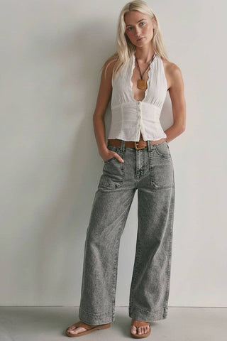 A woman modeling a white sleeveless button-front top and wide-leg gray jeans with a brown belt, standing against a plain light gray wall.
