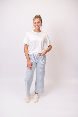 A woman modeling a white short-sleeve t-shirt and light blue and white vertically striped wide-leg cropped pants, paired with white sneakers, against a plain white studio background.