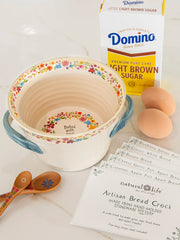 A beige stoneware hand molded bread crock with a floral pattern, a blue lid, and some decorative eggs and a carton of Domino sugar in the background, indicating preparation for baking.