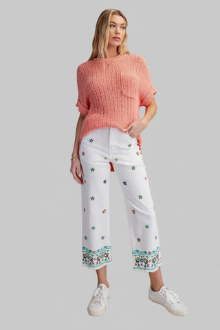 Woman wearing a pink sweater and white pants with floral embroidery on a beige background